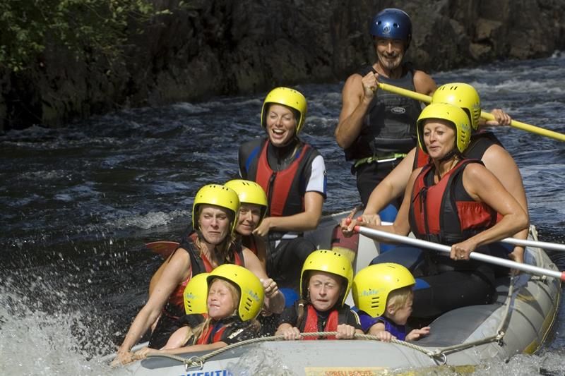 Rafting mit Menschen in schwarzen Neoprenanzügen und roten Schwimmwesten + gelben Helmen.