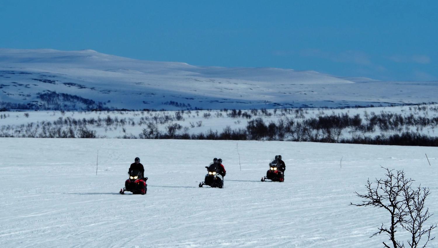 Trois motoneiges circulent sur un vaste champ de neige sous un ciel bleu.
