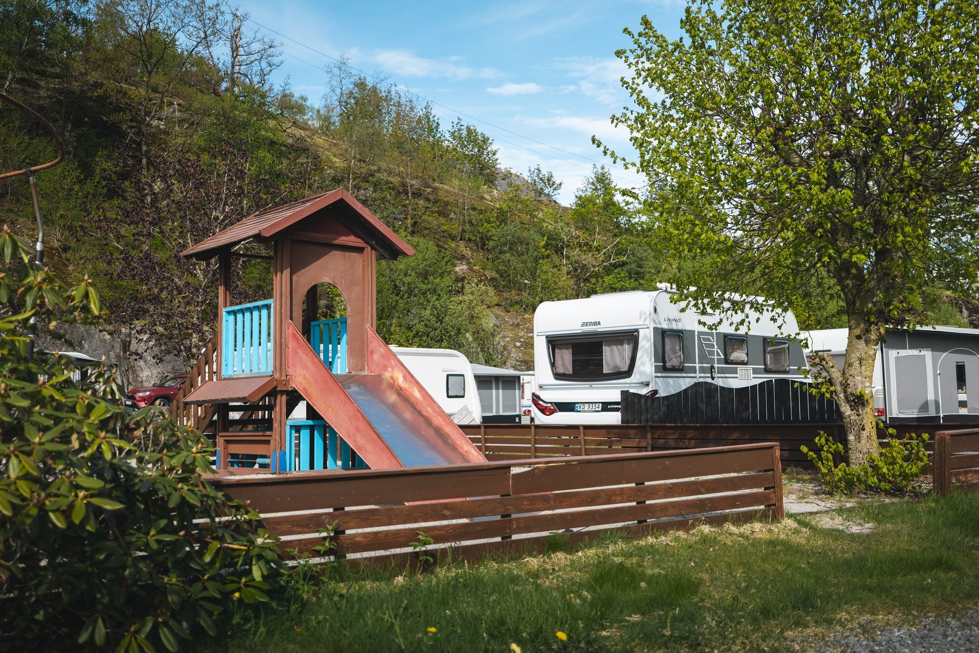 Wooden playground tower with a slide beside parked caravans, surrounded by trees and a rocky hillside under blue sky..