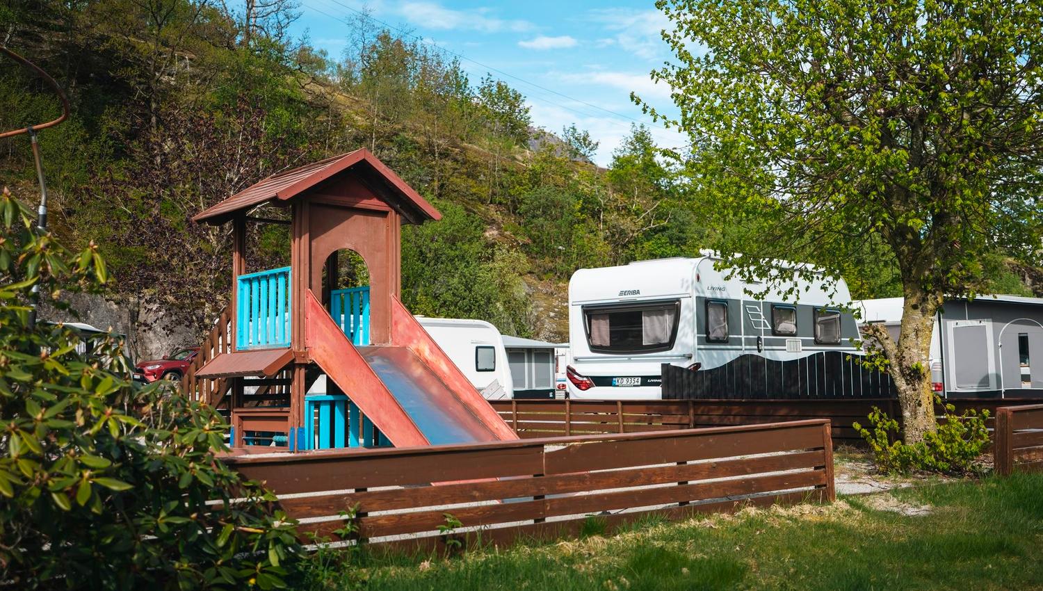Wooden playground tower with a slide beside parked caravans, surrounded by trees and a rocky hillside under blue sky..