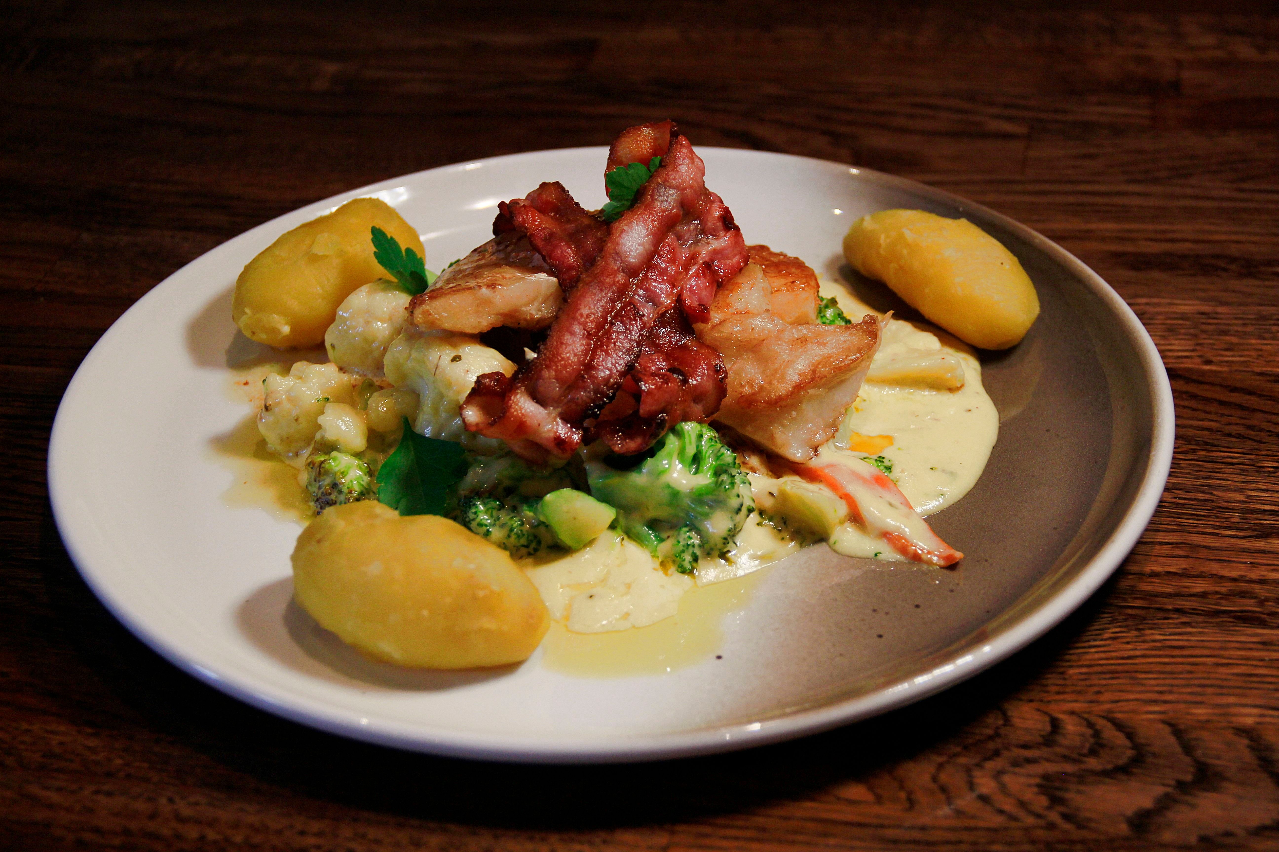 Fish with bacon on top and vegetables underneath, white sauce and three potatoes on a white plate