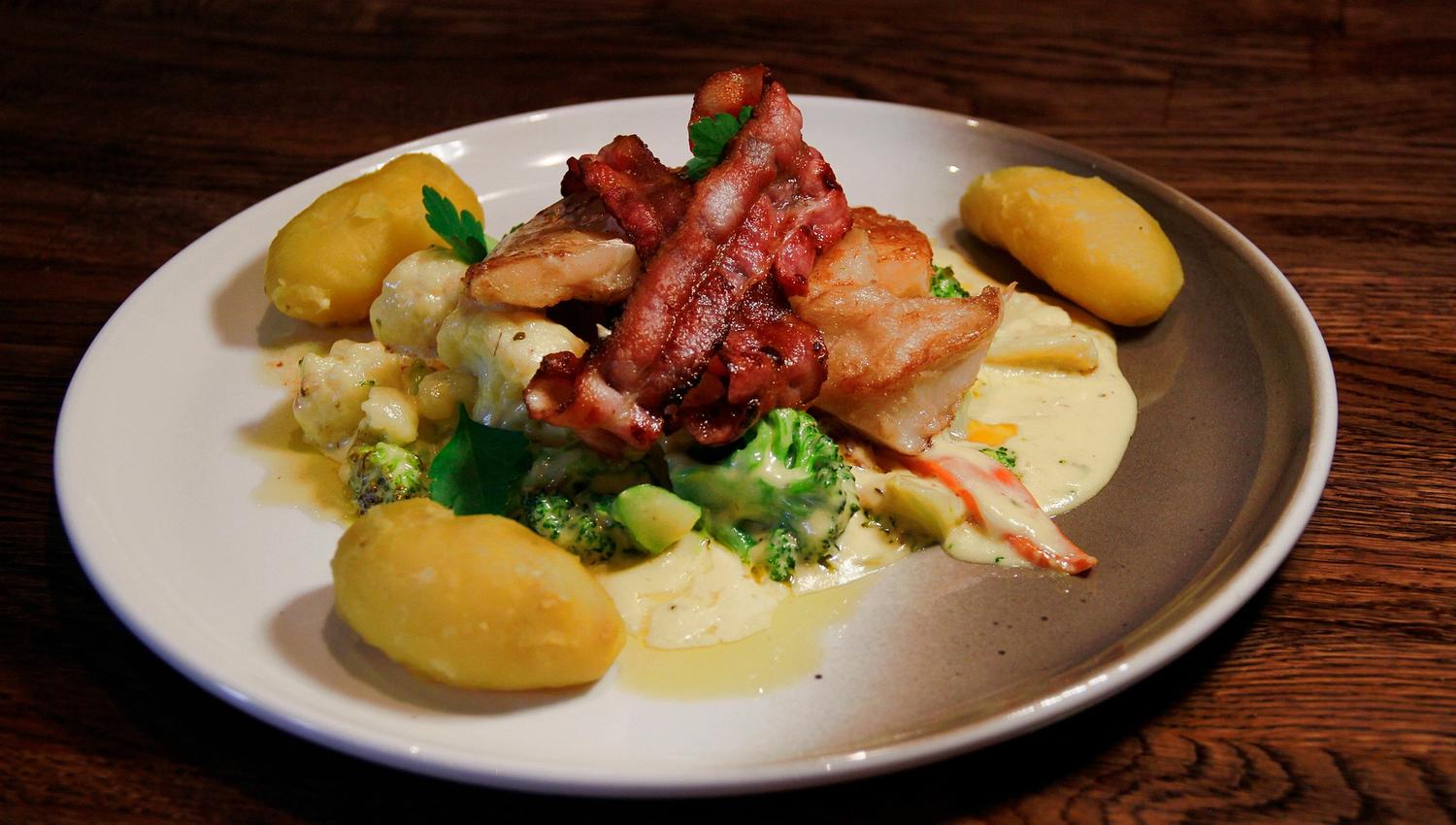 Fish with bacon on top and vegetables underneath, white sauce and three potatoes on a white plate