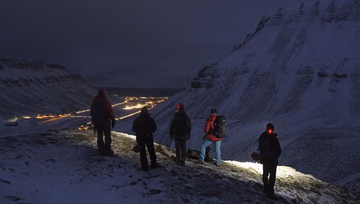 En gruppe med gjester som bruker hodelykter på tur i mørke omgivelser, med lys fra Longyearbyen i bakgrunnen