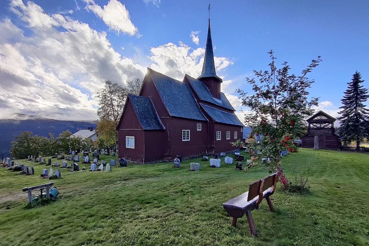 Stavkirke på grønn plen med et lite rognebærtre i motsol.