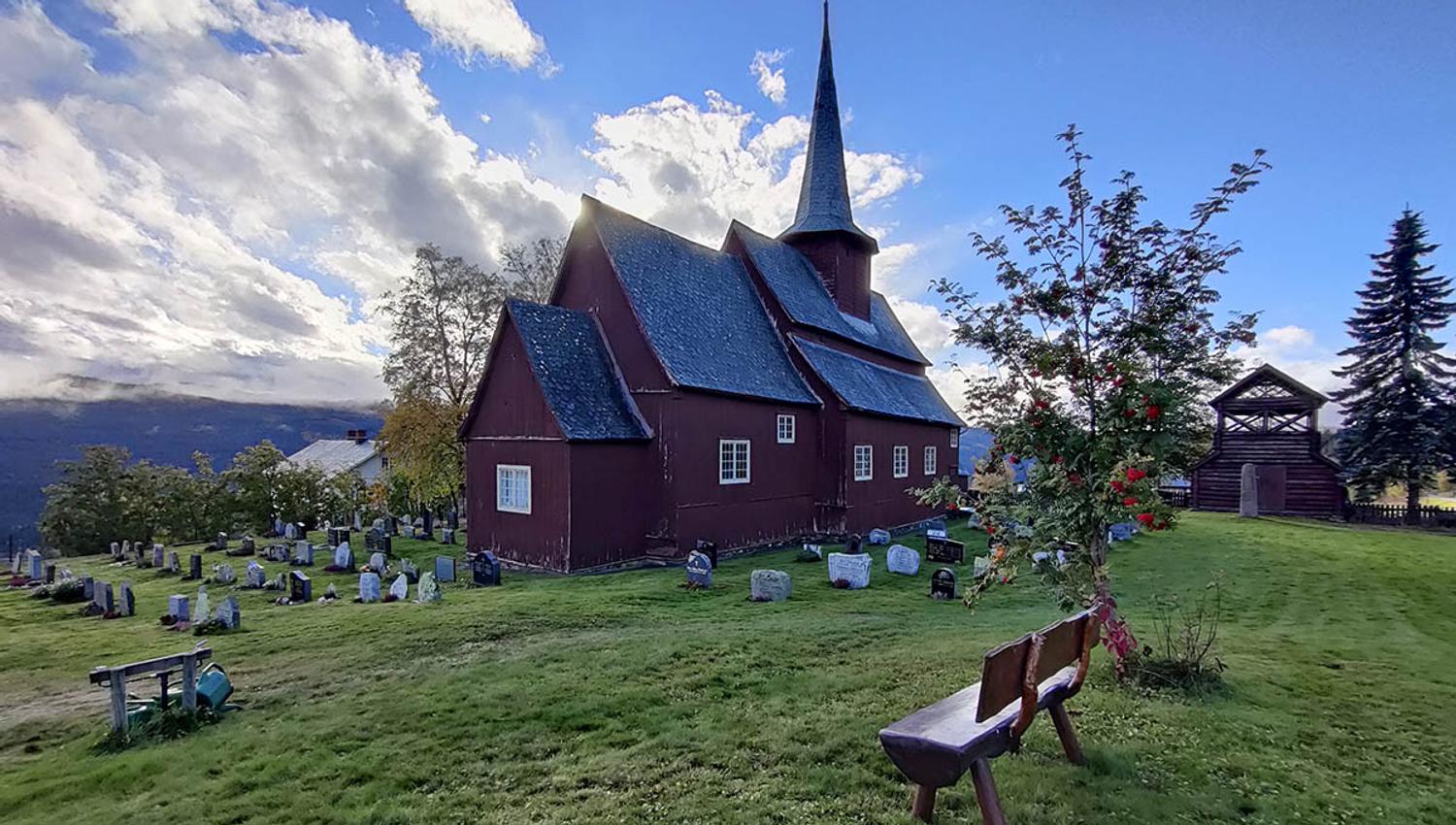 Stavkirke på grønn plen med et lite rognebærtre i motsol.