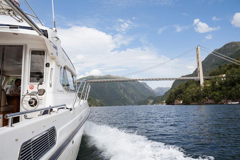 Hardanger Fjordcruise under an impressive bridge in the Hardangerfjord, surrounded by mountains and a blue sky.