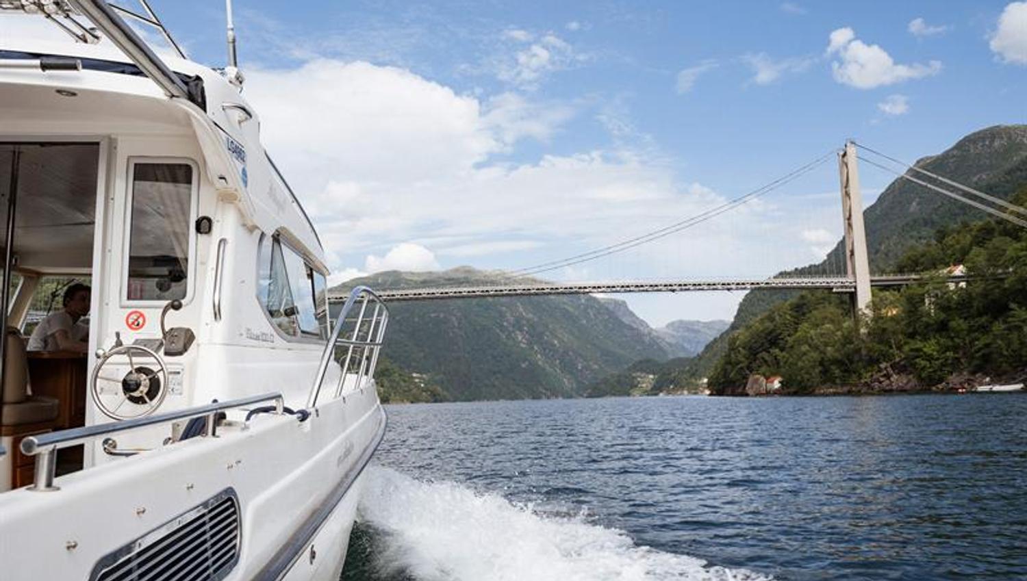 Hardanger Fjordcruise under an impressive bridge in the Hardangerfjord, surrounded by mountains and a blue sky.