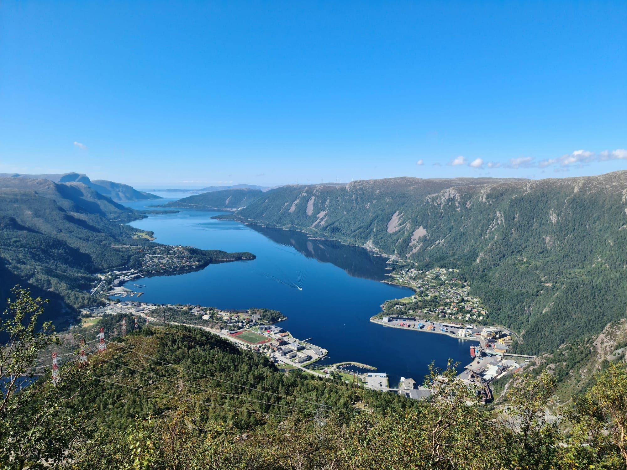 Panoramautsikt frå fjell over ein blå fjord med bygder, hamn og grøne fjellsider under klar himmel..