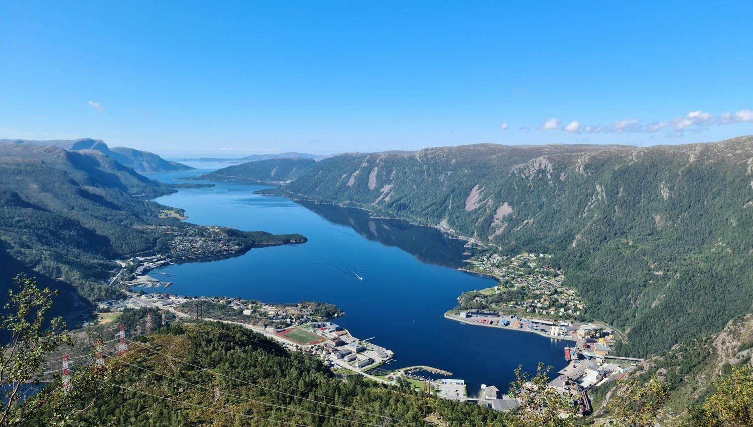 Panoramautsikt frå fjell over ein blå fjord med bygder, hamn og grøne fjellsider under klar himmel..