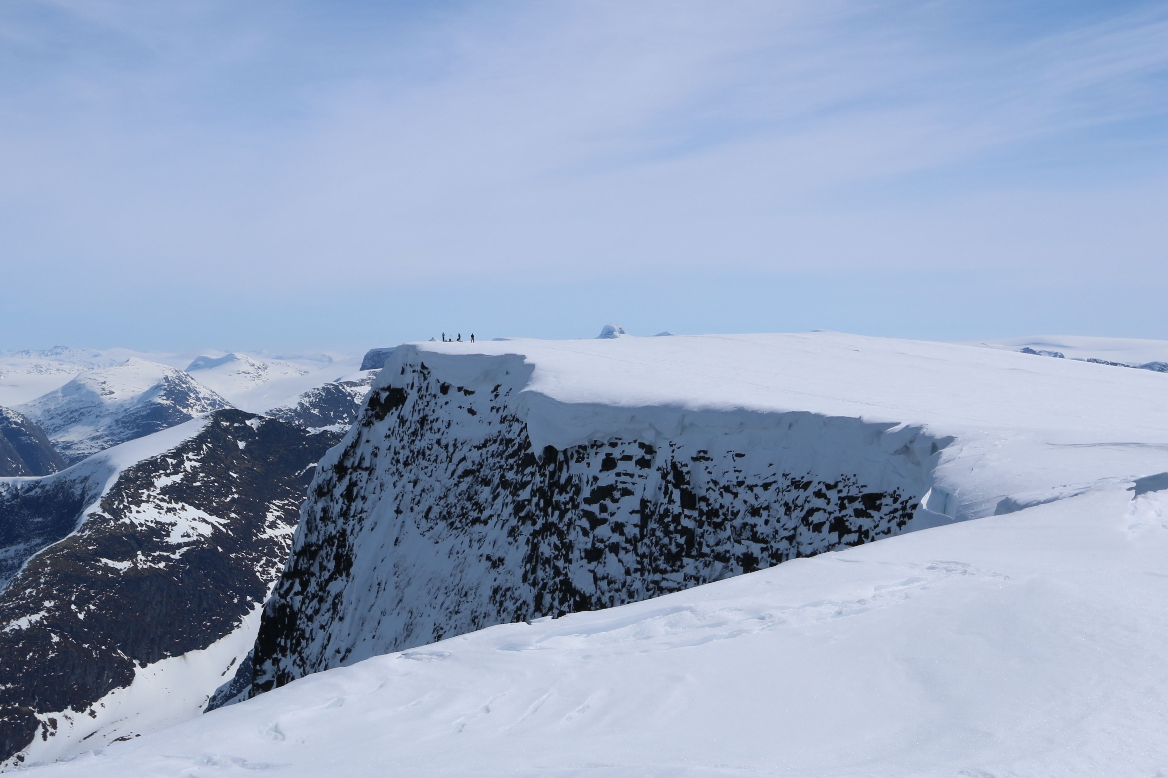 Wide view of a snow-covered mountain plateau with a steep rocky cliff and tiny hikers on the ridge under blue sky.