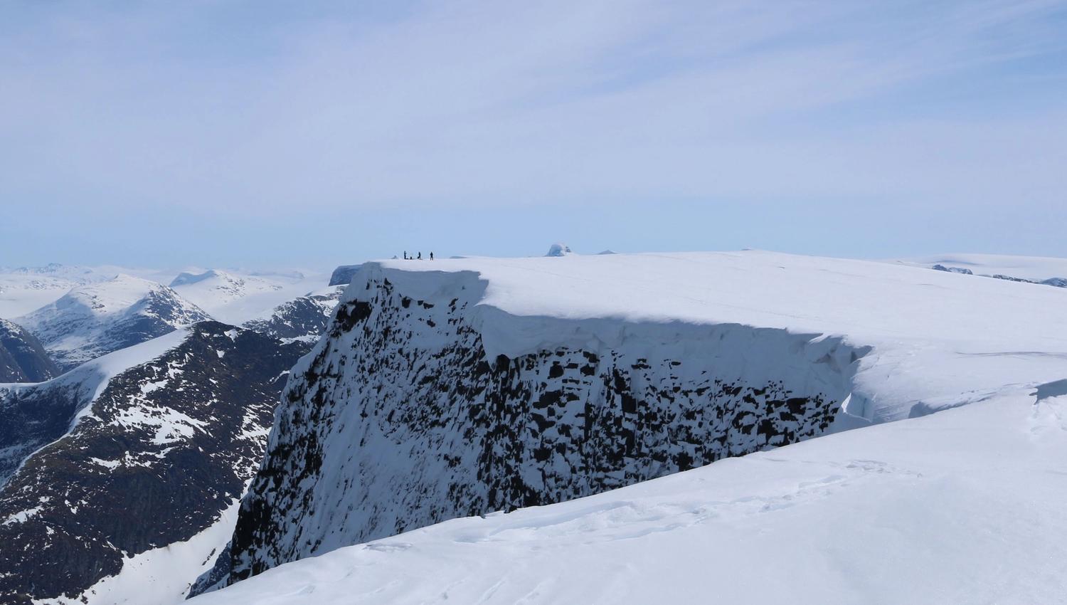 Wide view of a snow-covered mountain plateau with a steep rocky cliff and tiny hikers on the ridge under blue sky.