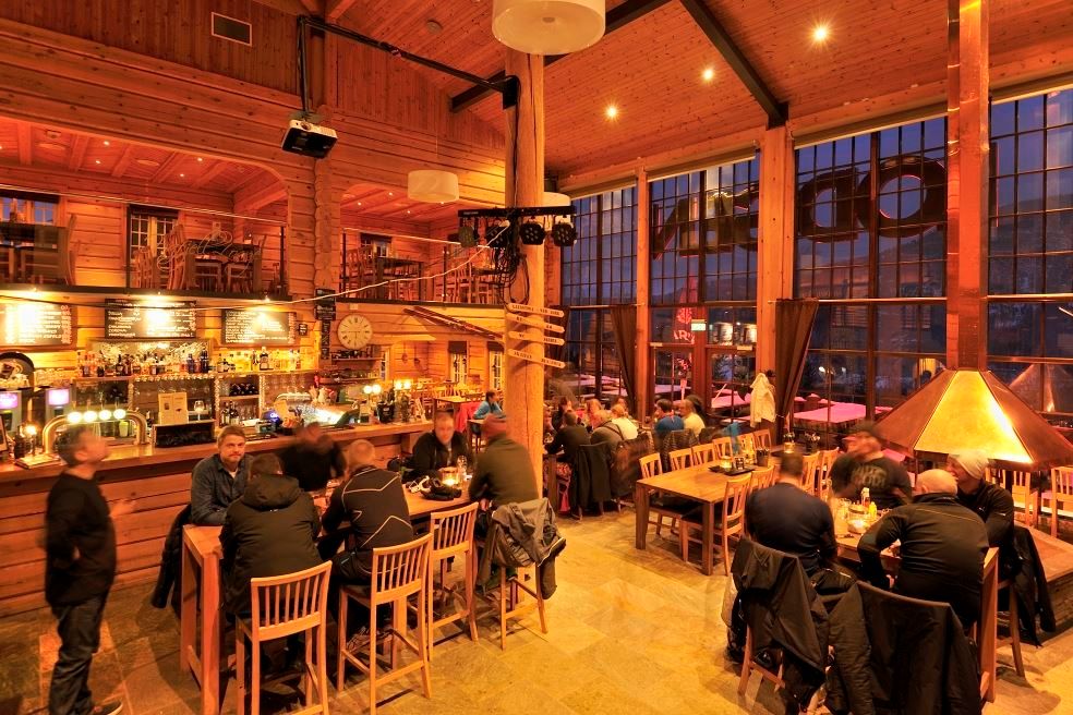 Rustikk bar i tre med mange gjester ved bord og bardisk, varm belysning og store vinduer mot kvelden..