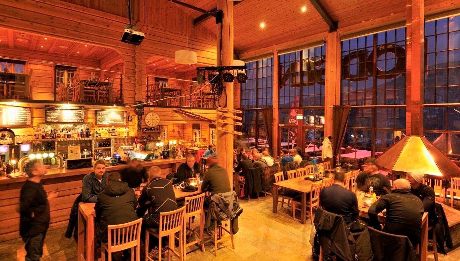 Rustikk bar i tre med mange gjester ved bord og bardisk, varm belysning og store vinduer mot kvelden..