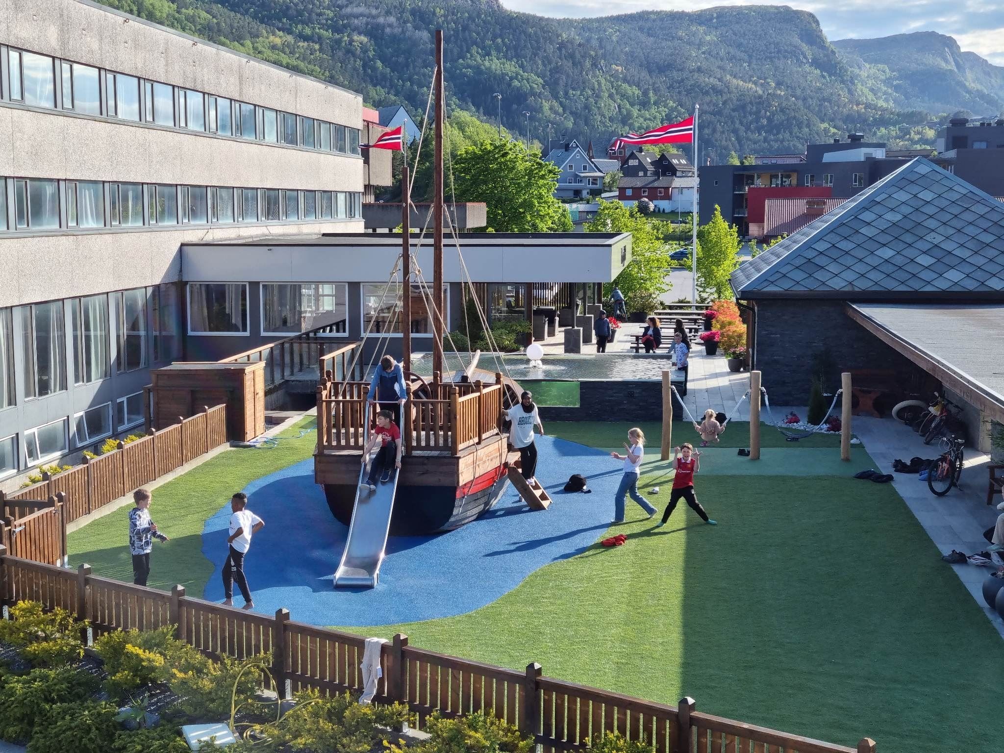 Born leikar utanfor Bremanger Fjordhotell. med piratskipleikeapparat og sklie, med fjell og bygningar i bakgrunnen..