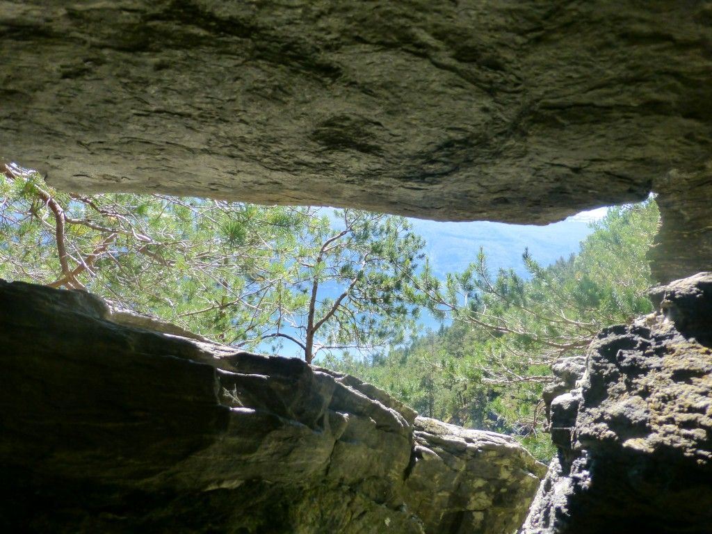 View from beneath a rocky overhang looking out to pine trees and distant blue mountains in daylight.