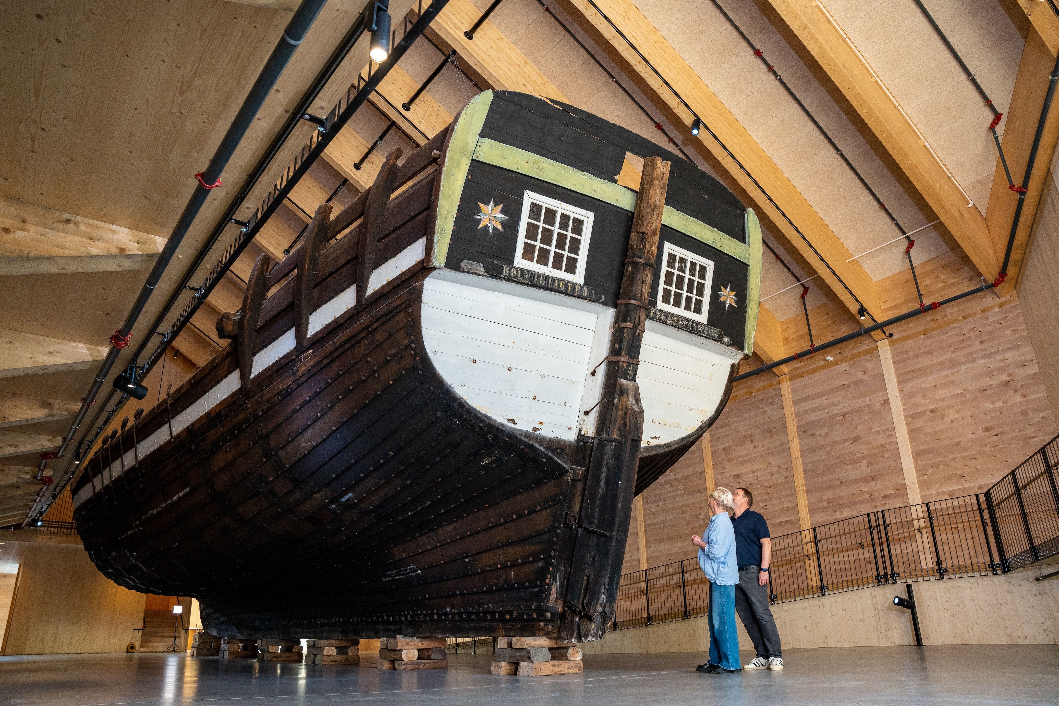 Large wooden ship Holvikejekta hull elevated on blocks inside a timber-framed hall, with two visitors standing nearby..