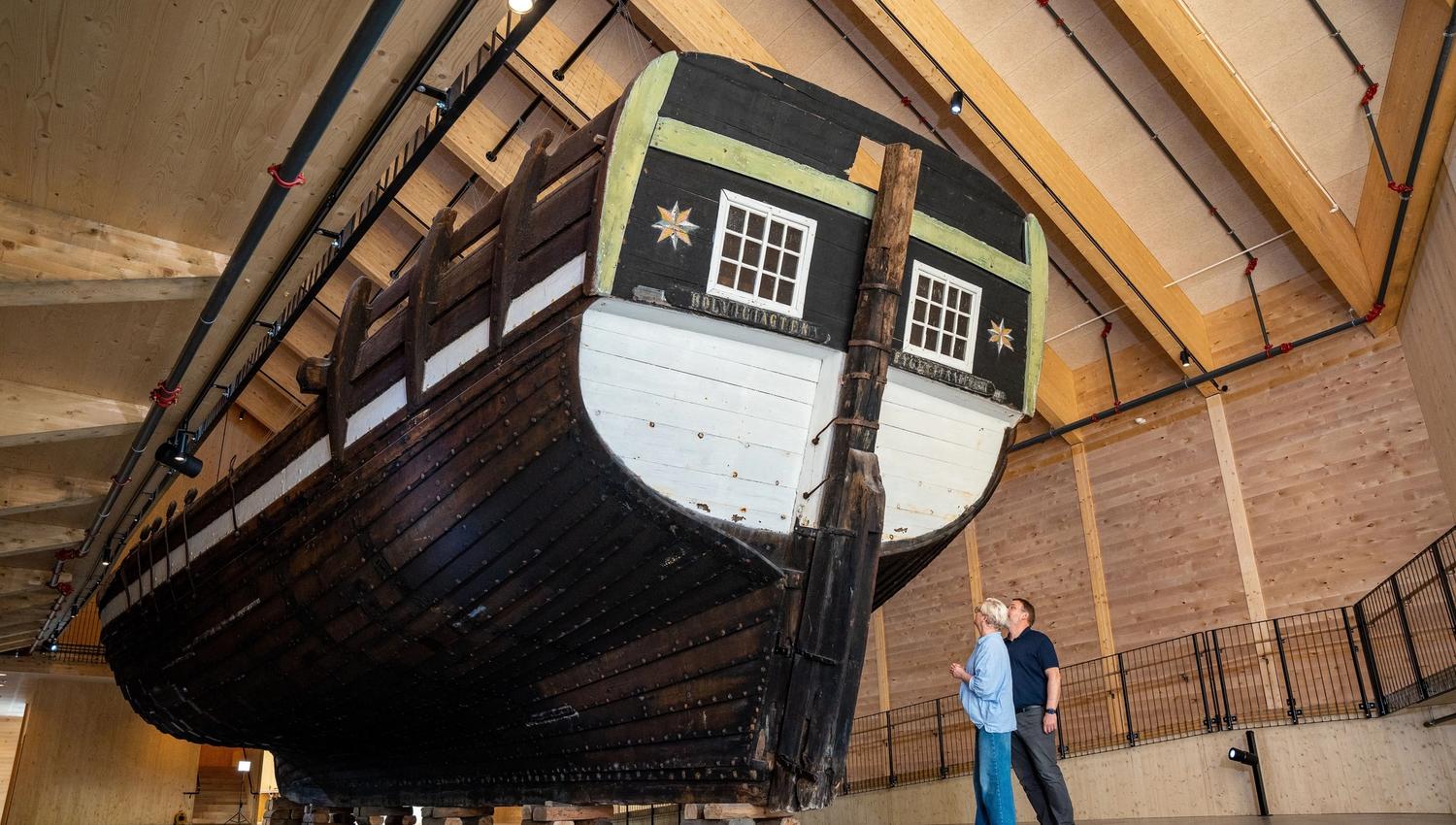 Large wooden ship Holvikejekta hull elevated on blocks inside a timber-framed hall, with two visitors standing nearby..
