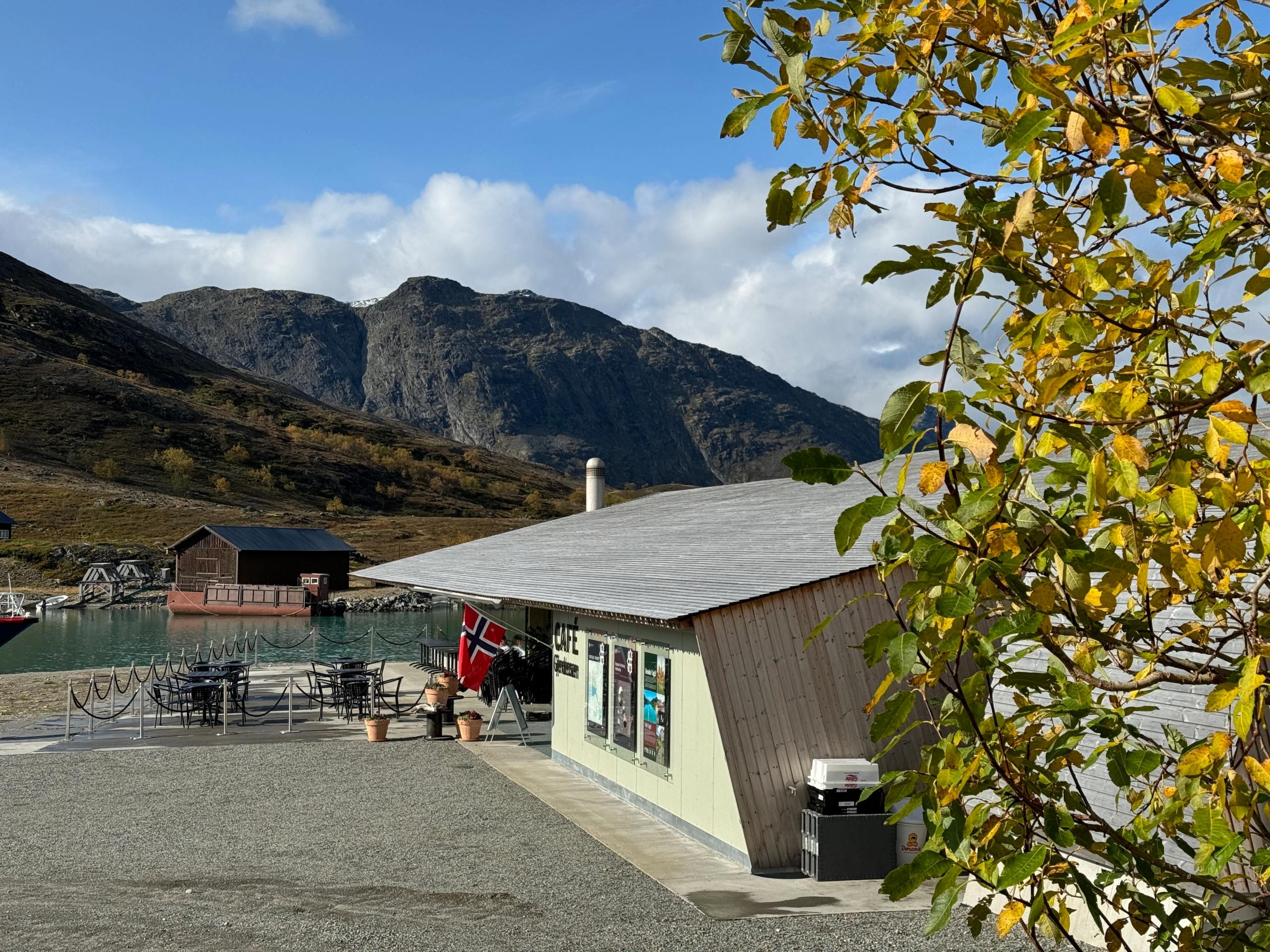 A lakeside cafe with outdoor tables and a Norwegian flag, set against rugged mountains and a leafy tree..