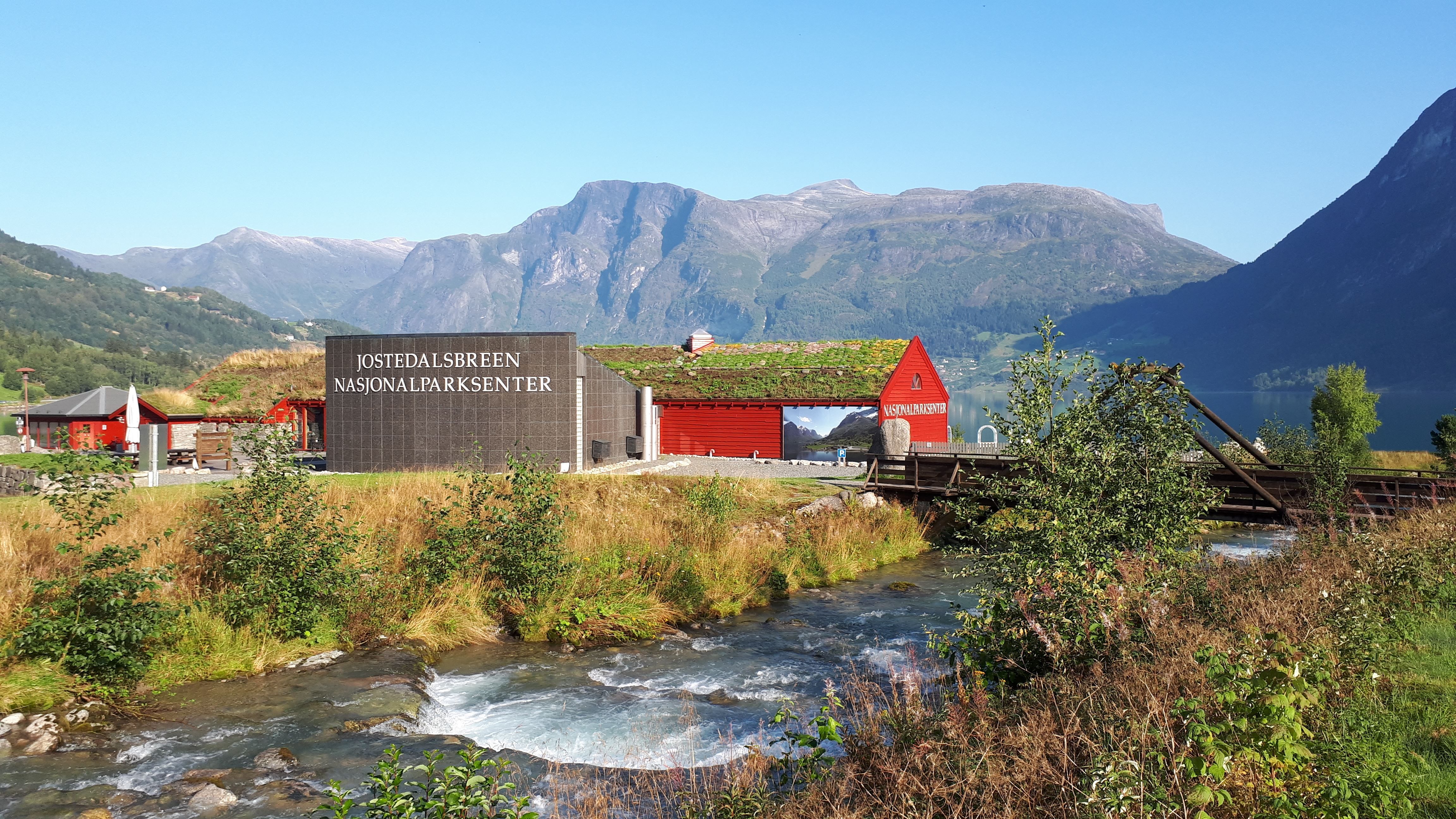 Raude bygg ved Jostedalsbreen nasjonalparksenter, med elv i forgrunnen og fjell og fjord under klar blå himmel..