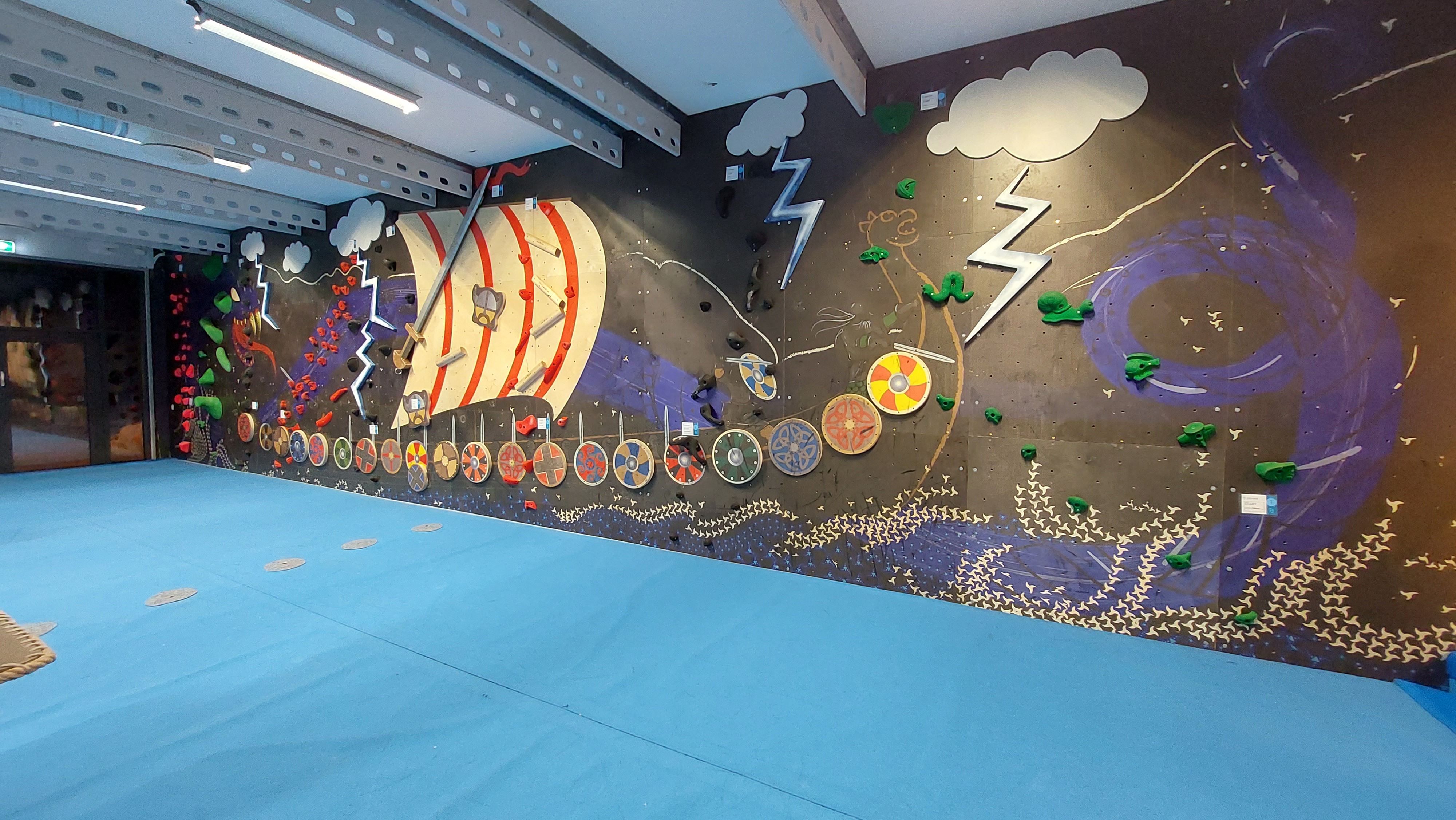 Indoor bouldering wall with a painted Viking ship mural, lightning clouds, green holds, and blue padded flooring..