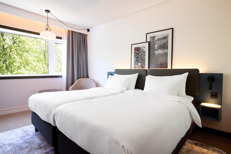 Lyst hotellrom med to enkeltsenger, store vinduer og moderne innredning.