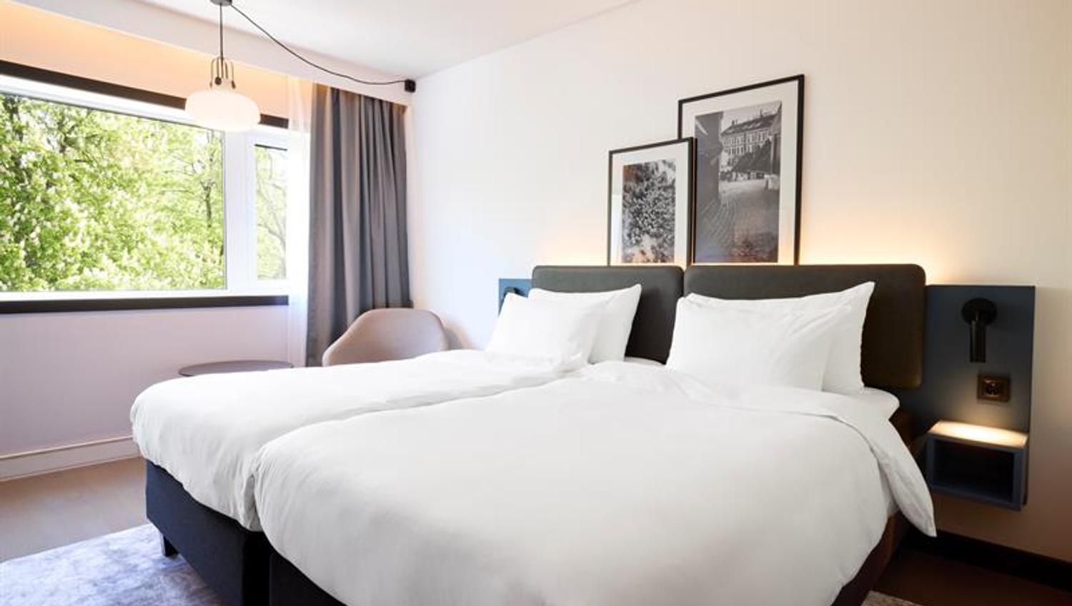 Lyst hotellrom med to enkeltsenger, store vinduer og moderne innredning.
