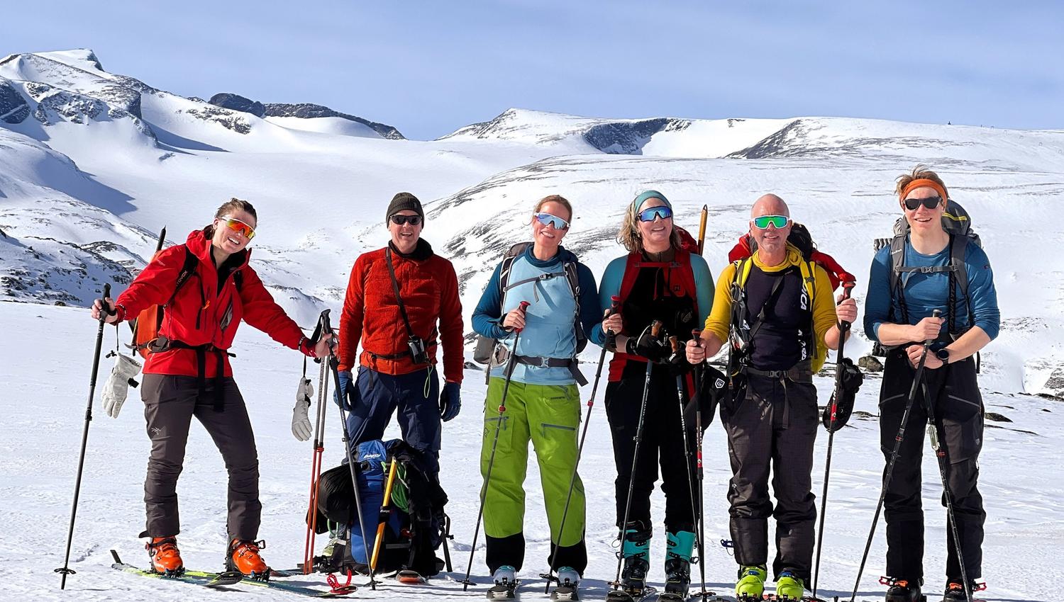 Zes skiërs met rugzakken en skistokken staan op een besneeuwde kam met heldere alpine bergen onder een heldere lucht..