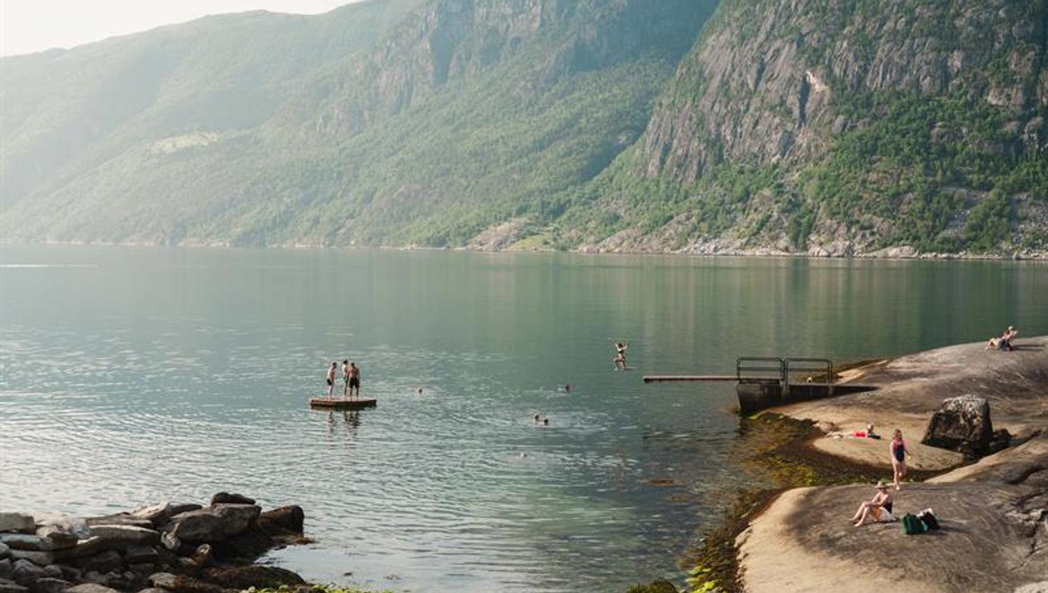 Kråkeskarvet beach with a person jumping from the diving board into the fjord. There are a lot of people sunbathing and swimming.