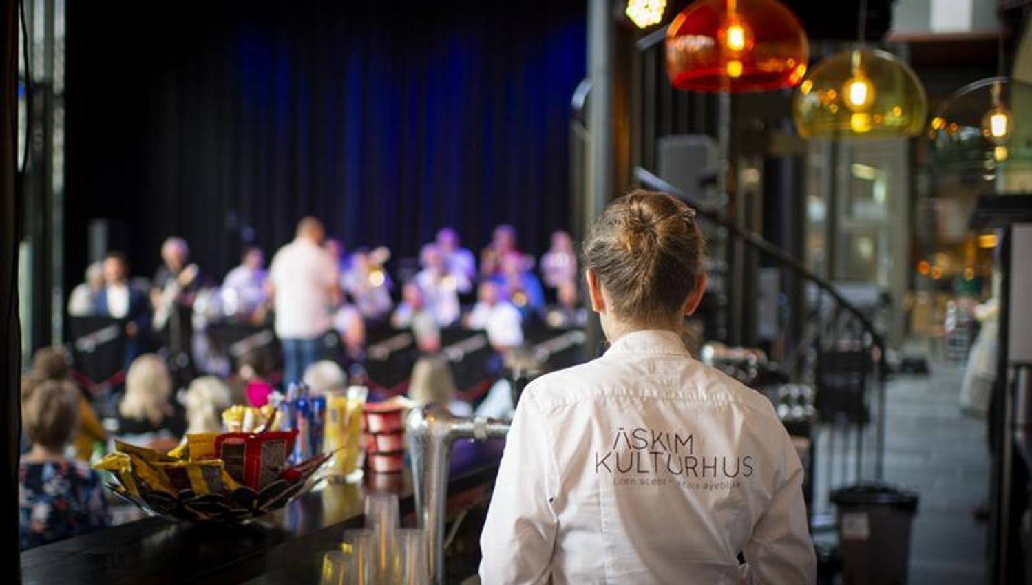 A person wearing a shirt labeled “Askim Cultural Center” stands by a bar counter, watching a band perform on stage. The audience is seated and listeni