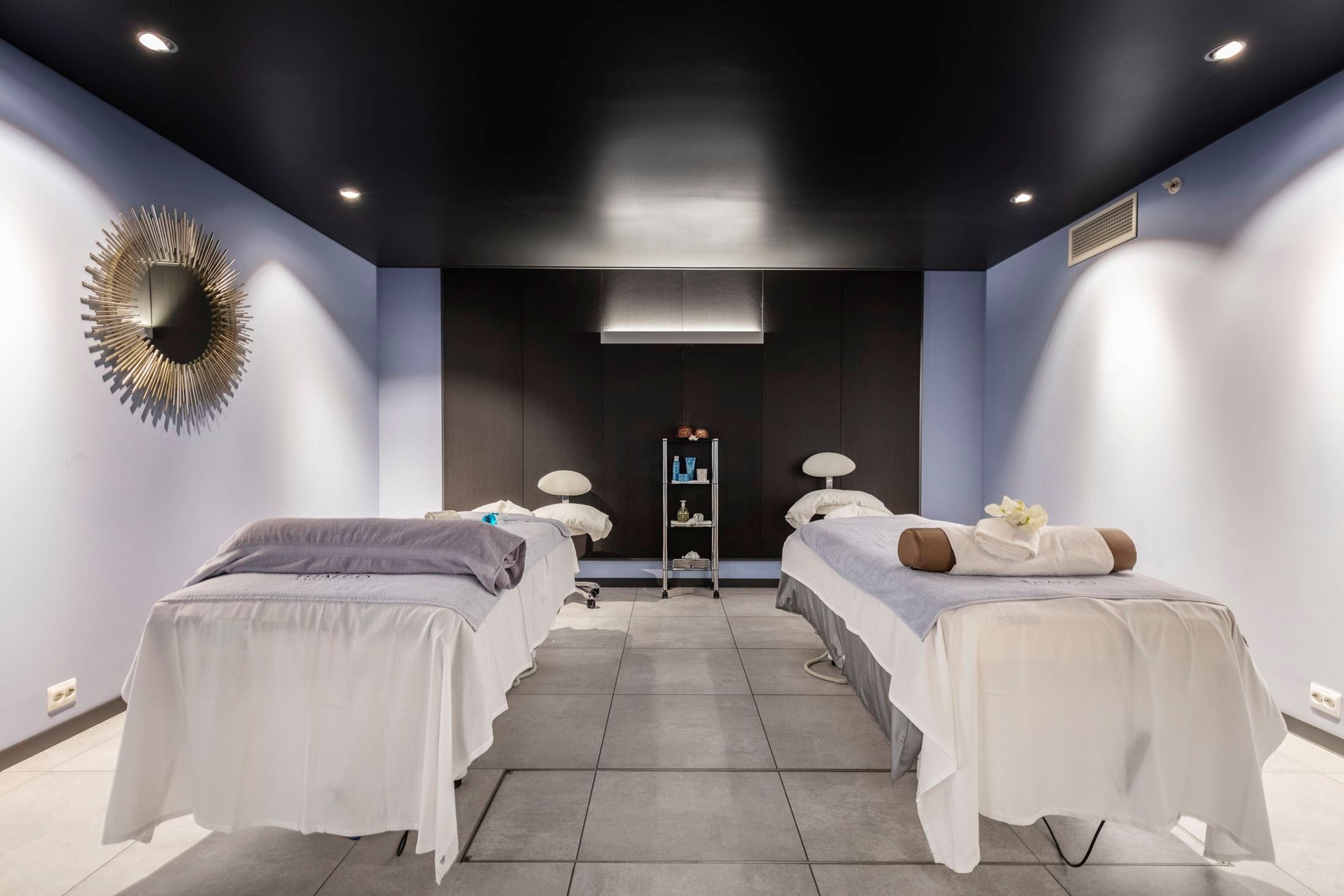 A modern spa room with two massage tables, folded towels, a small supply cart, and recessed ceiling lights at Alexandra Spa..