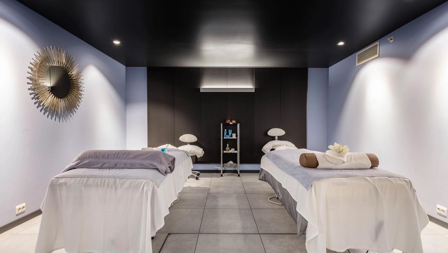 A modern spa room with two massage tables, folded towels, a small supply cart, and recessed ceiling lights at Alexandra Spa..