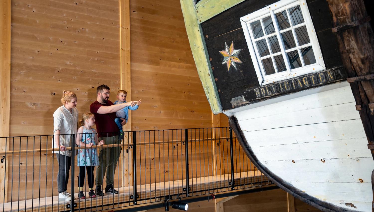A group of visitors stands on an indoor walkway beside a large wooden boat hull, Holvikejekta, with a window and painted star..