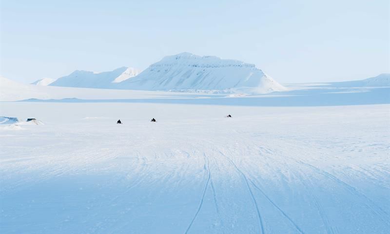 Snøscootere ses i det fjerne, i et åpent og snødekt landskap. Fjell i bakgrunnen