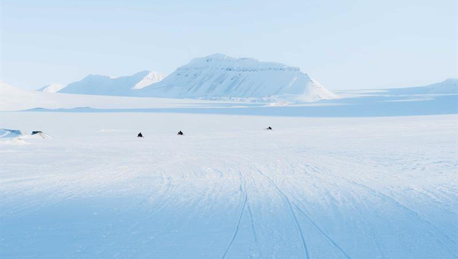Snøscootere ses i det fjerne, i et åpent og snødekt landskap. Fjell i bakgrunnen