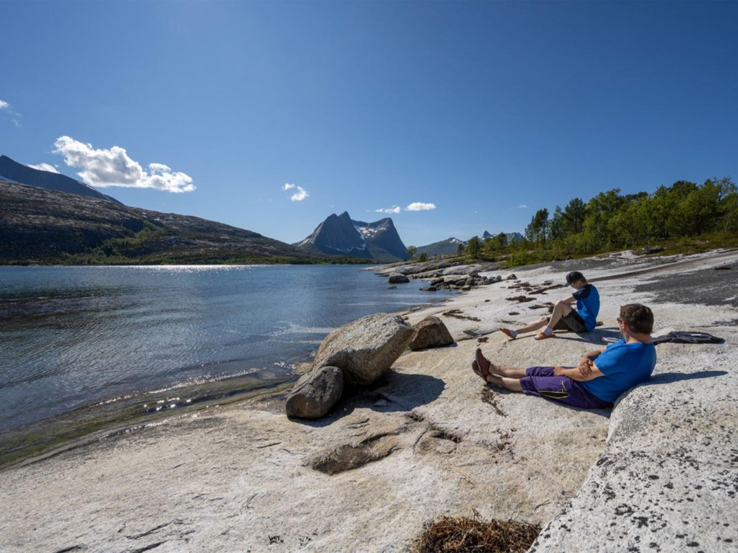 Two people relax on a rocky lakeshore beside calm water, with distant sharp mountains under a clear blue sky.