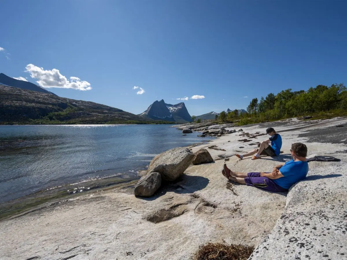 Two people relax on a rocky lakeshore beside calm water, with distant sharp mountains under a clear blue sky.