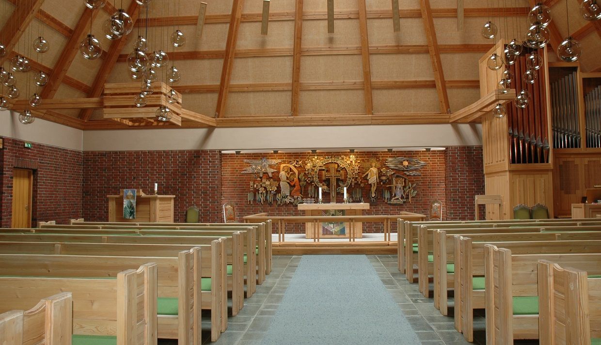 The church from the inside. Wooden benches with green pillows. Chandelairs hanging from the ceiling. A large mural behind the alter.