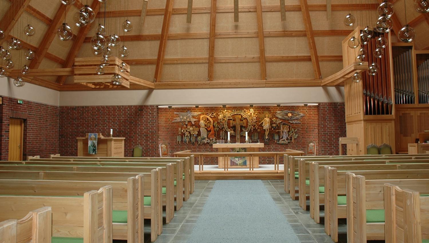The church from the inside. Wooden benches with green pillows. Chandelairs hanging from the ceiling. A large mural behind the alter.
