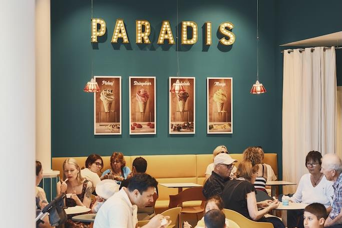 Mennesker som sitter ved små, runde bord og spiser inne i kaféen. Paradis-logoen på en grønn vegg bakerst i lokalet