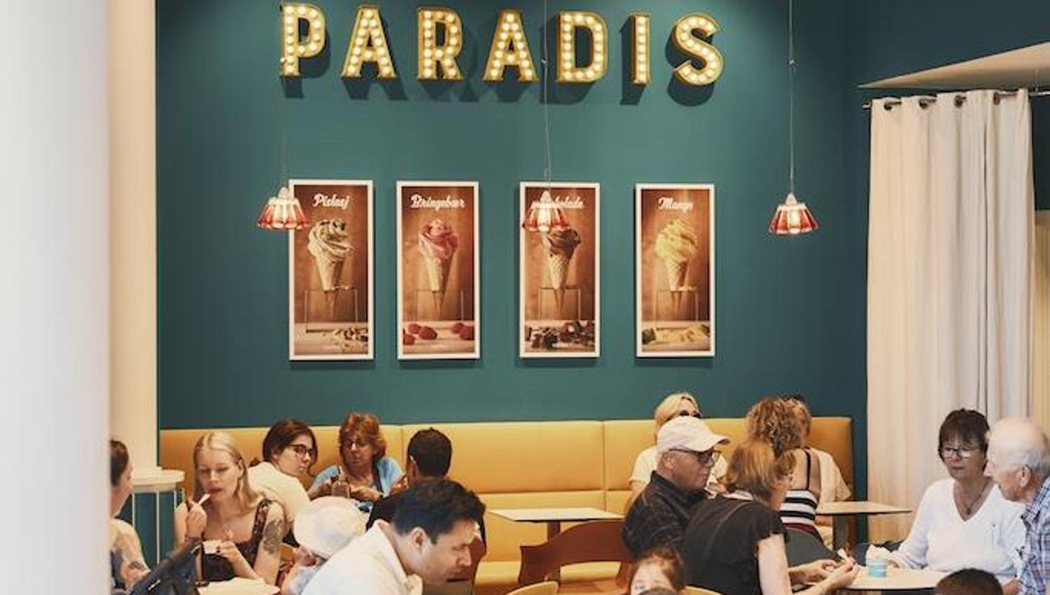 Mennesker som sitter ved små, runde bord og spiser inne i kaféen. Paradis-logoen på en grønn vegg bakerst i lokalet