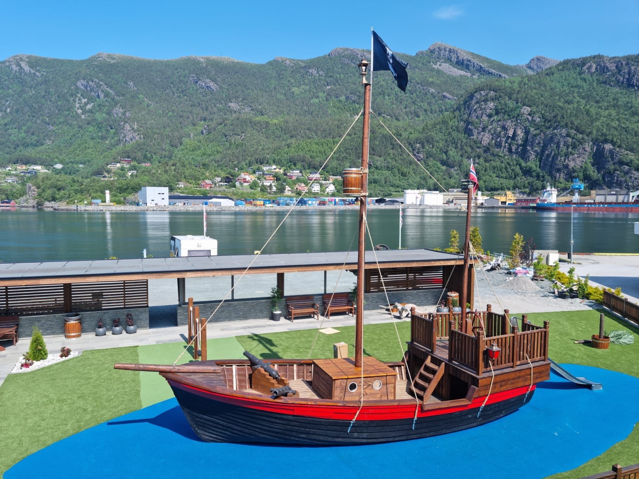 Ein dekorativ trebåt med mast står på grønt gras nær fjorden, med fjell og bygg på andre sida under blå himmel..