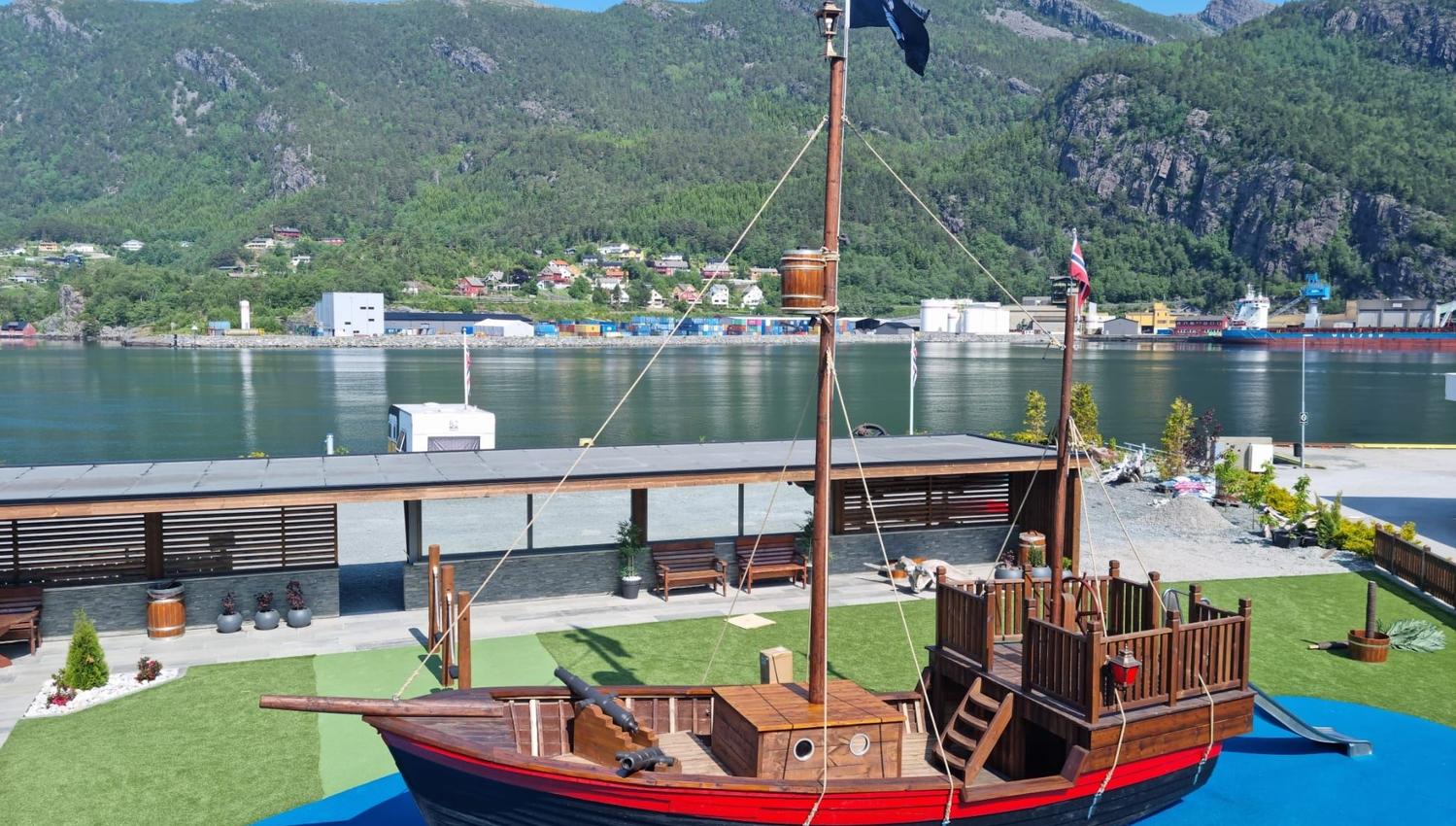 Ein dekorativ trebåt med mast står på grønt gras nær fjorden, med fjell og bygg på andre sida under blå himmel..