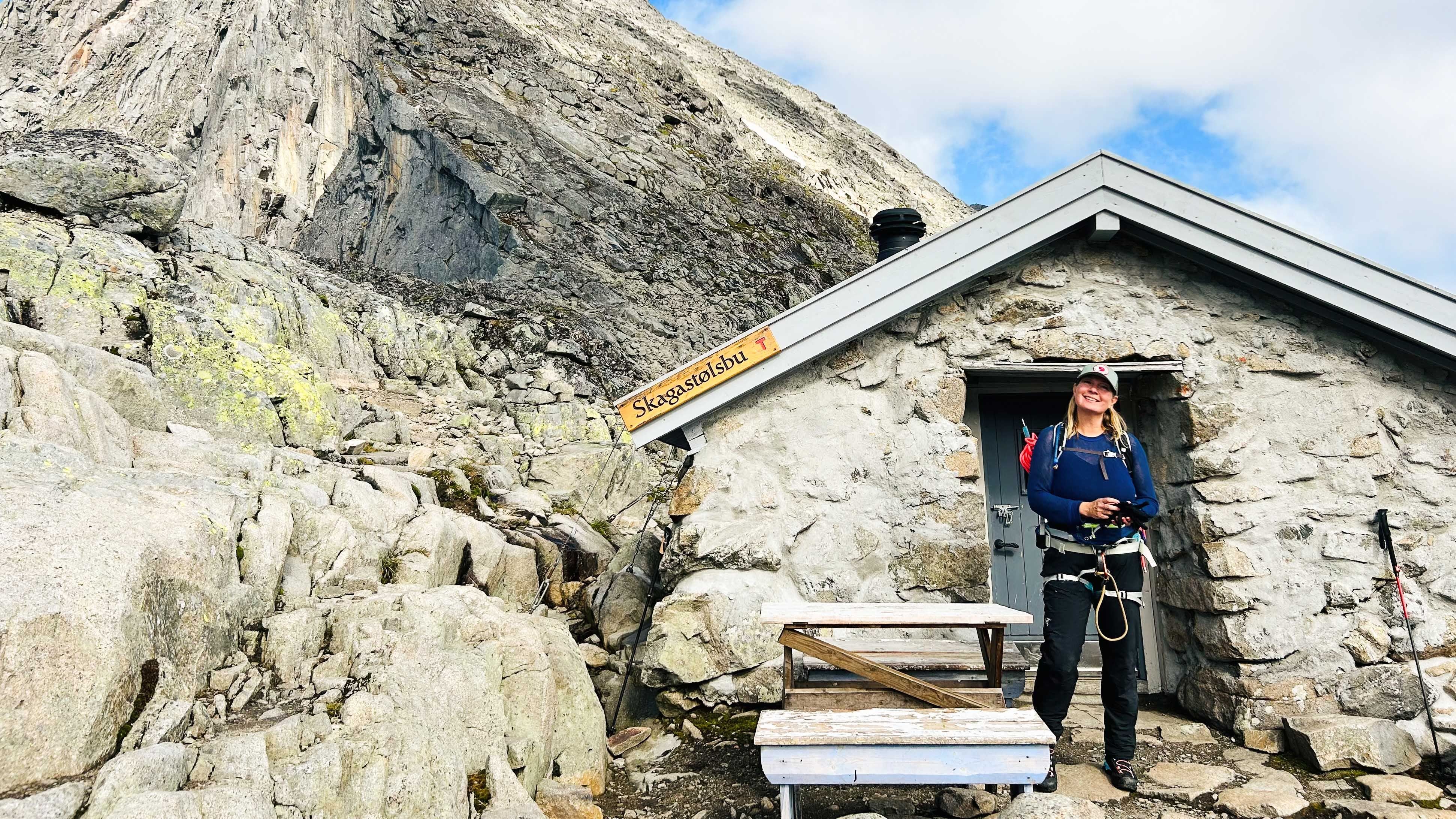 A hiker stands by the door of a small stone cabin on a rocky mountainside under a partly cloudy sky..