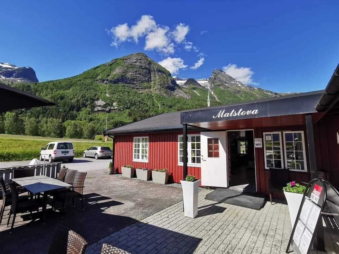 Red wooden café building with patio tables and parked cars, set beneath green mountains and a blue sky with clouds.