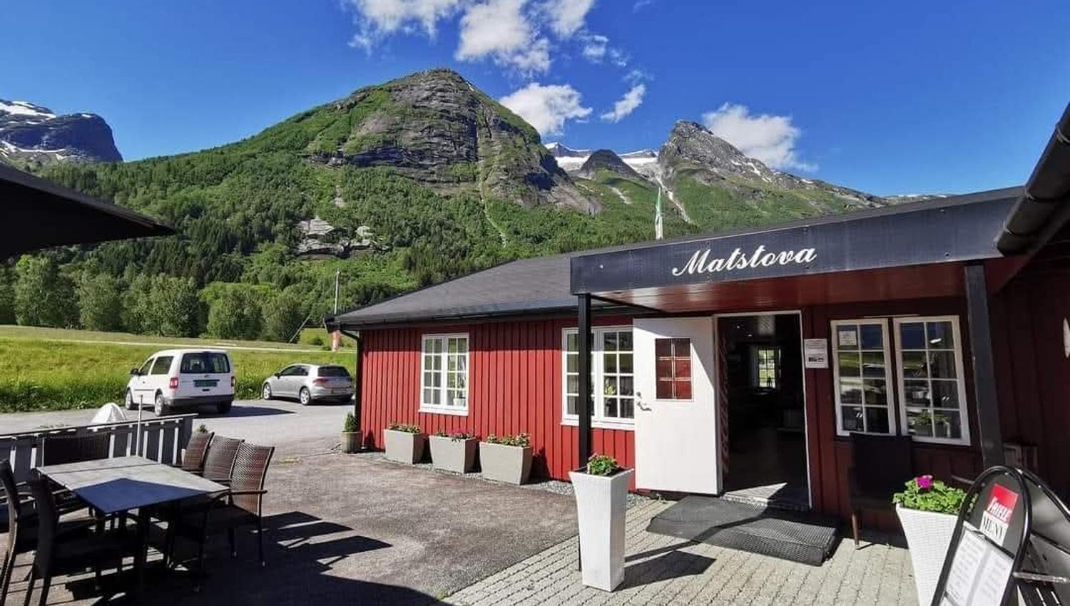 Red wooden café building with patio tables and parked cars, set beneath green mountains and a blue sky with clouds.