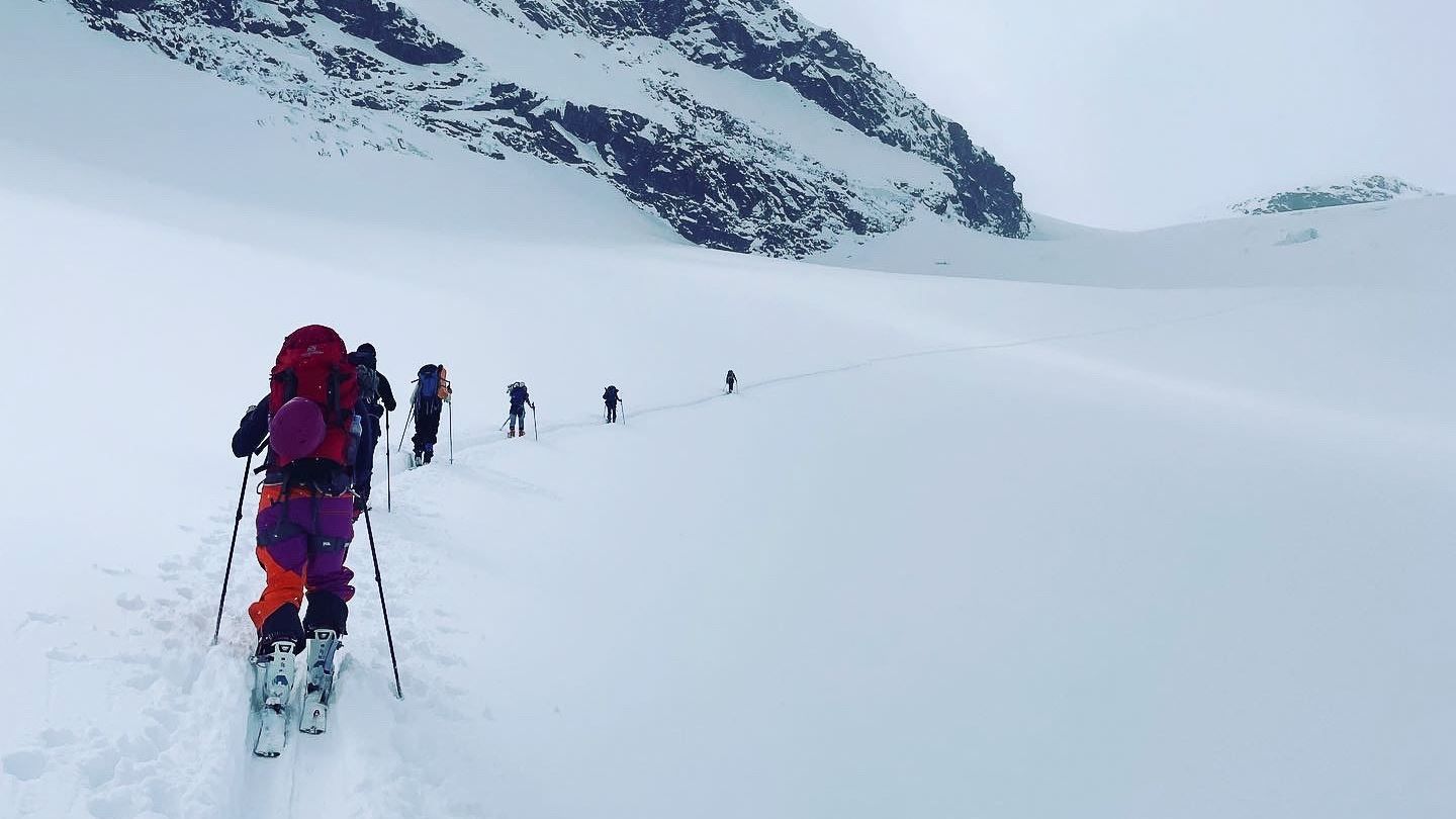 A line of skiers with backpacks and poles travels across a wide snowy slope beneath dark rocky mountains..