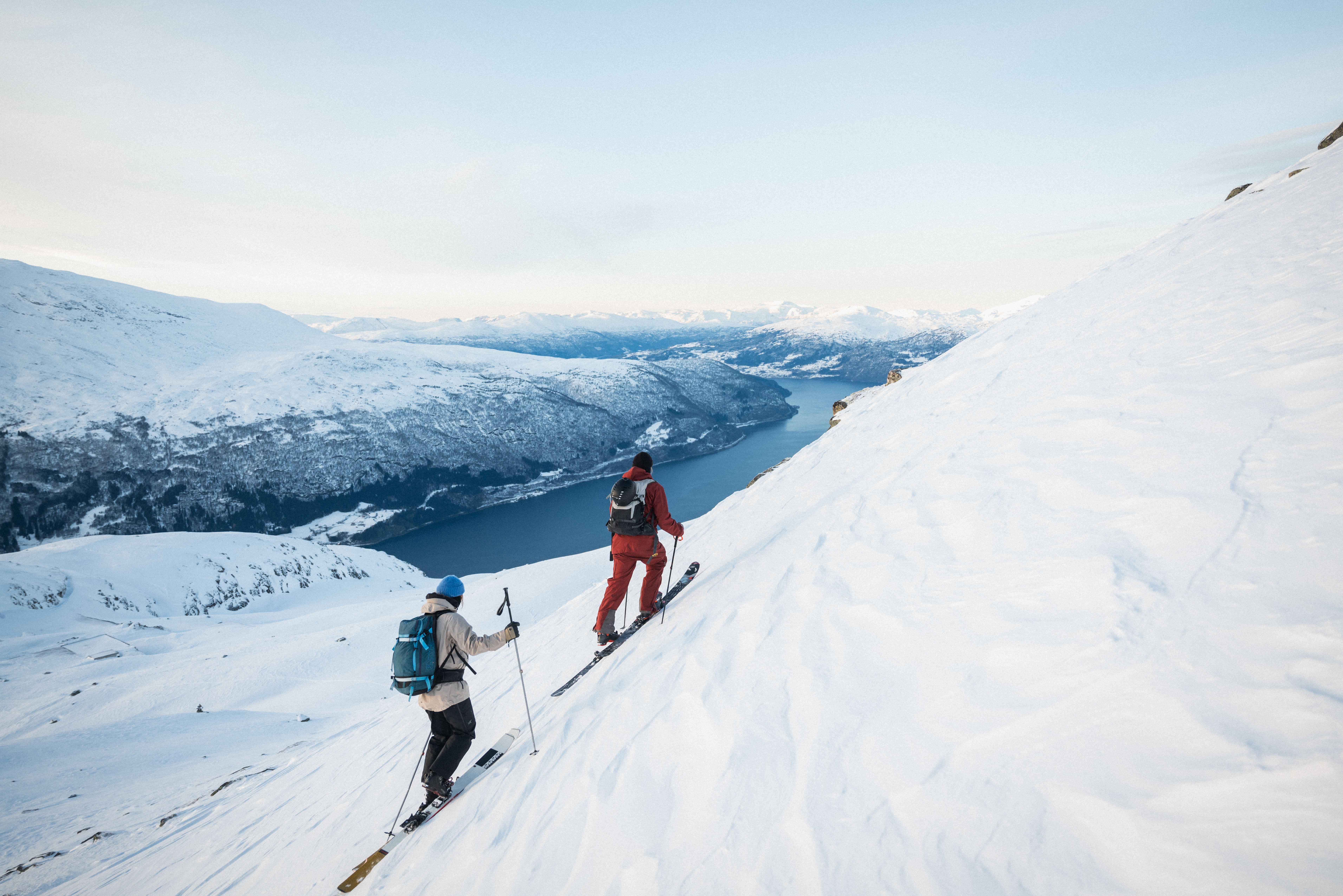 Two skiers climb a steep snowy mountainside with poles and backpacks, overlooking a fjord and distant peaks.