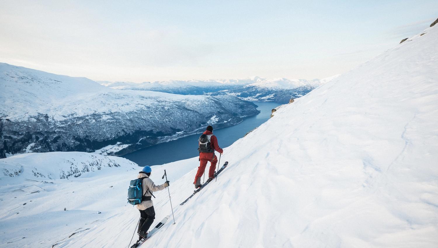 Two skiers climb a steep snowy mountainside with poles and backpacks, overlooking a fjord and distant peaks.