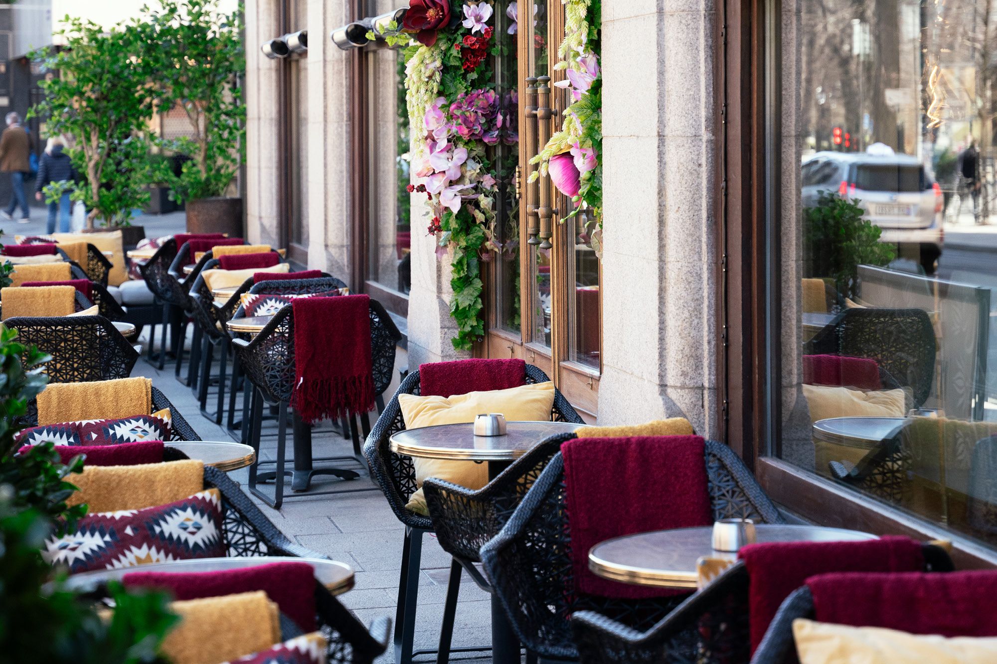 Outdoor seating with chairs and table with yellow pillows, ashtrays on the tables, flowers hanging on the wall.