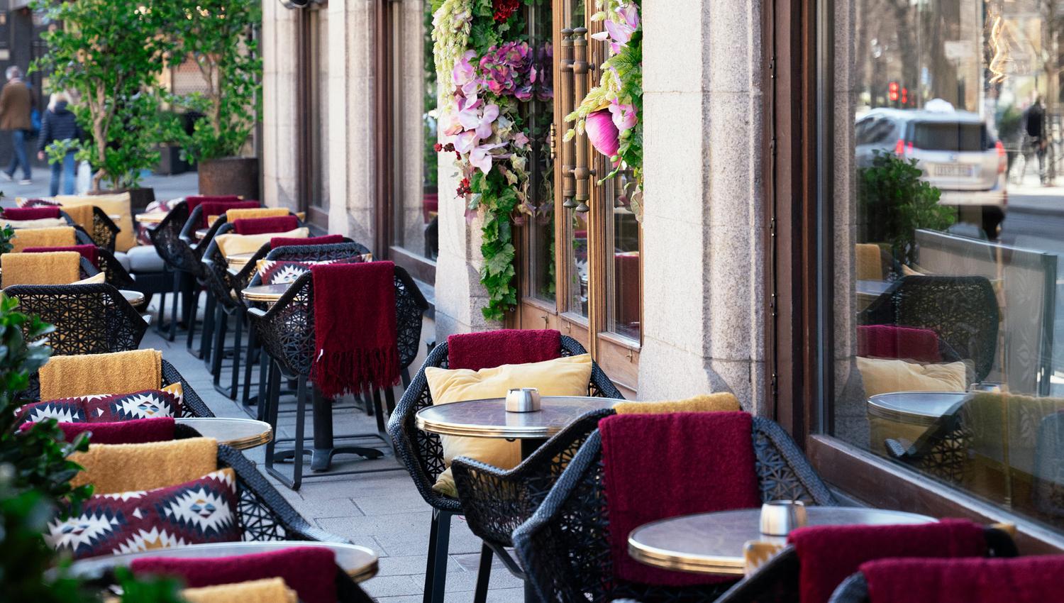Outdoor seating with chairs and table with yellow pillows, ashtrays on the tables, flowers hanging on the wall.